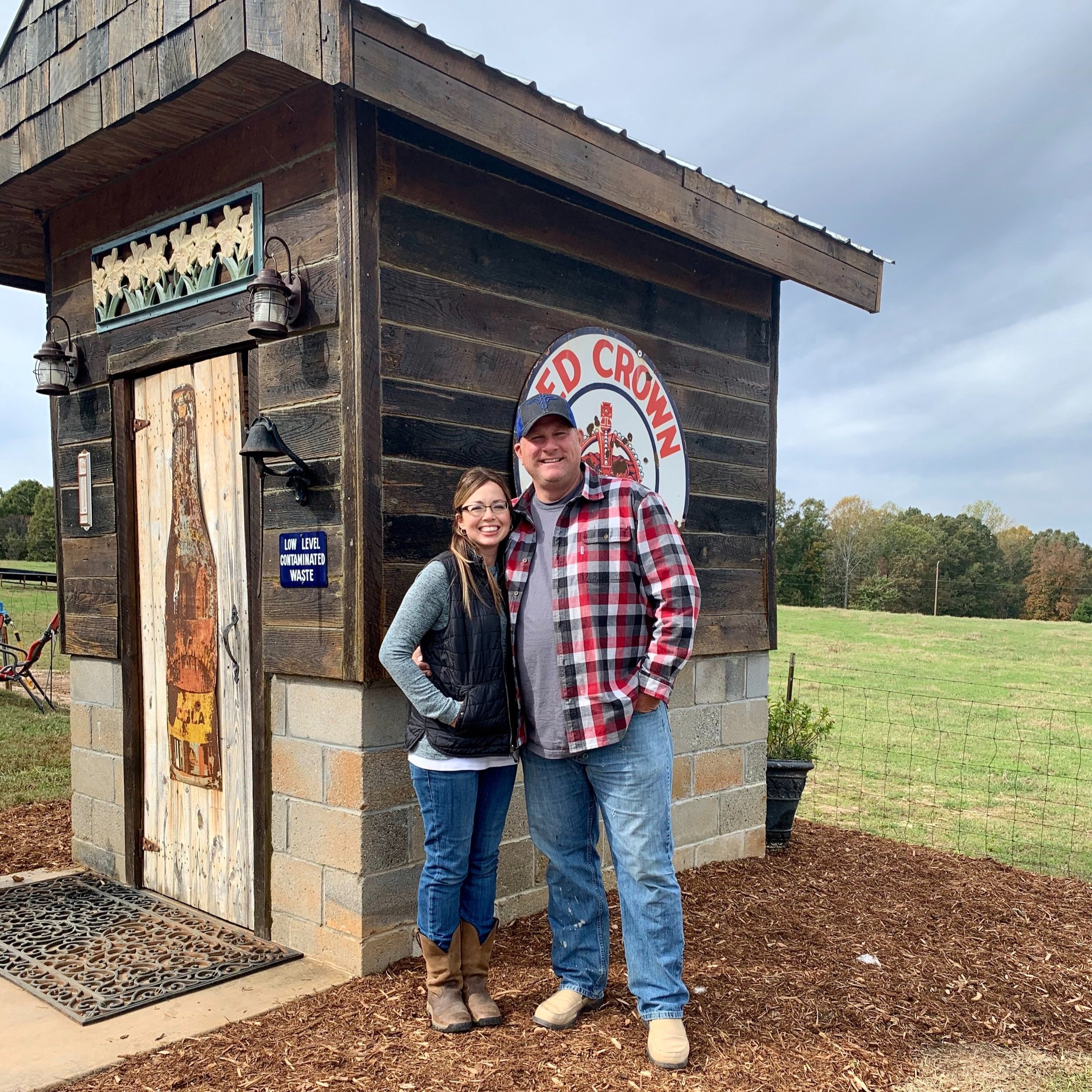Lukens+Land+&+Cattle+-+about+us+photo Couple posing in front of a rustic wooden building with a vintage soda sign, surrounded by a grassy field and trees, showcasing a charming outdoor setting.